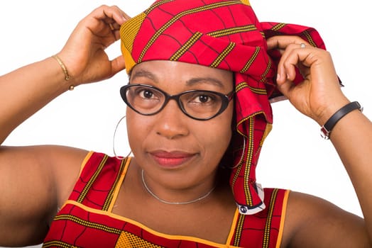 Close-up stylish smiling african mature woman in red african loincloth and glasses standing in studio and arranging her traditional headscarf on her head