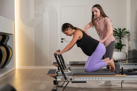 A pregnant woman works out on a reformer exercise machine with a personal trainer