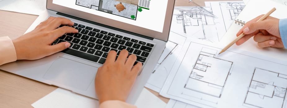 Eco house blueprint displayed on a laptop. Professional architect hand draws blueprint on meeting table with architectural document scatter around. Focus on hand. Closeup. Delineation.