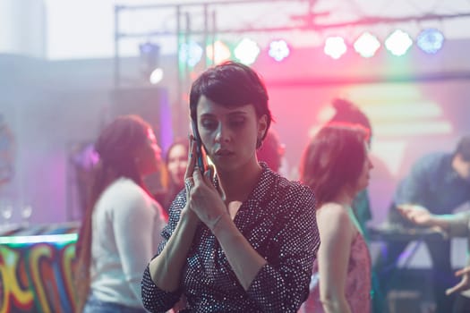 Young woman answering smartphone call while standing on dancefloor in nightclub. Serious clubber girl holding mobile phone and having communication while relaxing in club