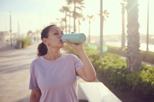 Wellness and healthy lifestyle. Young female athlete, attractive, fit, determined sportswoman drinking fresh clean water from a stainless steel bottle, taking a break after sports workout outdoors