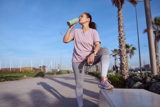 Sporty girl taking break after outdoor workout or morning jog, standing by a stone bench on the promenade and drinking refreshing water. Active people. Healthy lifestyle. Sport and fitness