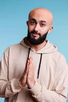 Arab man praying with hands folded in worship, expressing faith studio portrait. Young adult bald bearded person pleading, making wish and looking at camera with hopeful emotions