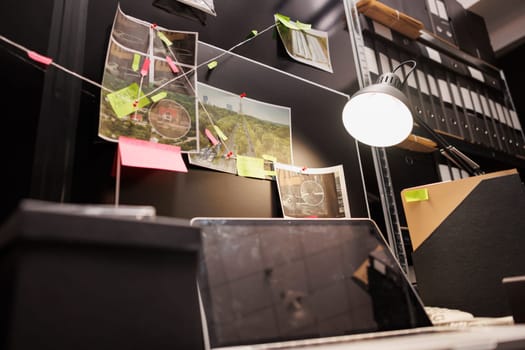 Crime scene photos attached to detective board over table in investigator room. Private detective workplace with case investigation map and laptop lit by desk lamp at night time