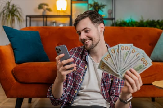 Successful rich young man holds smartphone and waving dollar bill money fan sitting in living room at home. Happy Caucasian guy winning online lottery game planning vacation calculating budget concept