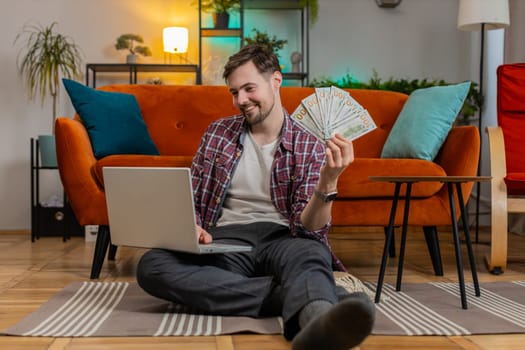 Planning budget. Rich happy Caucasian man counting money cash dollar using laptop calculate domestic bills at home. Young adult guy satisfied of income earnings, saves money for planned vacation