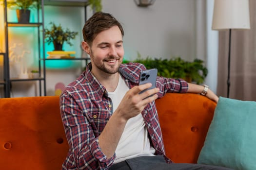Happy smiling man in casual clothes thinking and text messaging on smartphone. Caucasian guy sitting on sofa and using mobile phone in living room at home. Male is watching online movie in apartment.