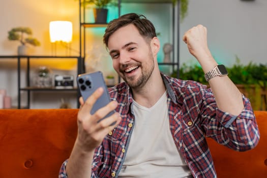 Surprised excited man winner holding smartphone reading good news amazed by online bet bid game win. Happy Caucasian guy on couch looking at screen overjoyed by lottery casino victory success at home