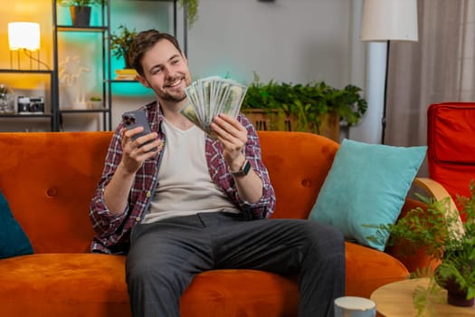 Successful rich man holding smartphone and waving dollar bill money fan sitting on orange sofa at home. Happy smiling young guy winning online casino lottery game planning vacation calculating budget