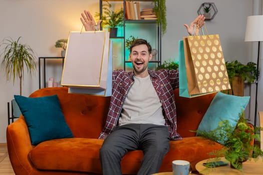 Young happy man shopaholic consumer came back home after shopping sale with bags. Discount sellout on mall amazed guy satisfied received parcels purchase from online order at home apartment on sofa