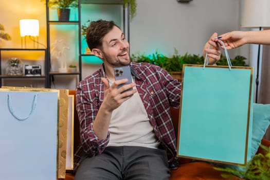 Man in casual clothes using smartphone and receiving shopping bags sitting on sofa in living room at home. Excited surprised Caucasian guy happy to receive online orders from courier. Discount sellout