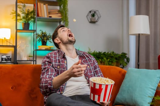 Man throwing eating popcorn watching interesting tv serial sport game online social media movie content sitting on sofa in room at home. Happy Caucasian guy in plaid shirt enjoying film during weekend