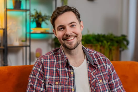 Portrait of happy Caucasian man sitting on sofa looking at camera and smiling at home. Young guy in plaid shirt having attractive appearance enjoying leisure time relaxing in living room in apartment