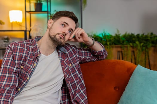Disappointed young man sits at home reflecting on moral distress after quarrel break-up or divorce problem feeling bad annoyed ill sick. Sad Caucasian guy in room contemplates the emotional aftermath.