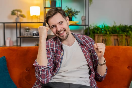 Happy Caucasian man fingers crossed watching TV shouting, celebrating success, winning, goal achievement good news, lottery luck, victory sits on sofa in living room. Guy clenching fists at home