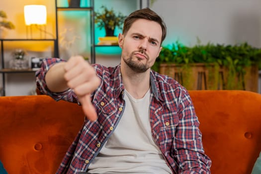 Dislike. Upset unhappy young man showing thumbs down sign gesture, expressing discontent, disapproval dissatisfied bad work at home apartment indoors. Displeased angry Caucasian guy sitting on sofa.