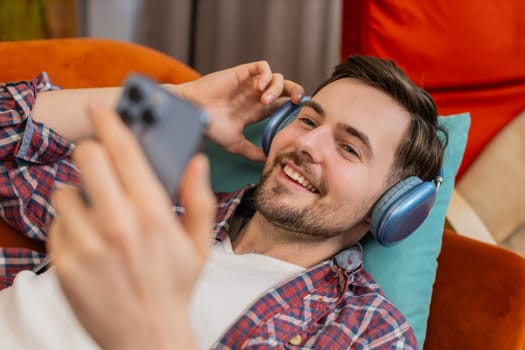 Happy Caucasian young man in wireless headphones choosing listening favorite energetic dancing music on smartphone. Smiling guy relaxing on sofa in living room at home in apartment during weekends