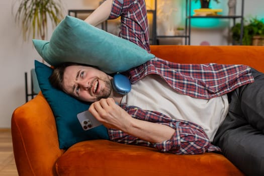 Repair work at neighbors. Irritated angry man lying on sofa covers ears with cushions annoyed by noisy loud music suffer from headache wishes silence. Thin walls at home flat without sound insulation