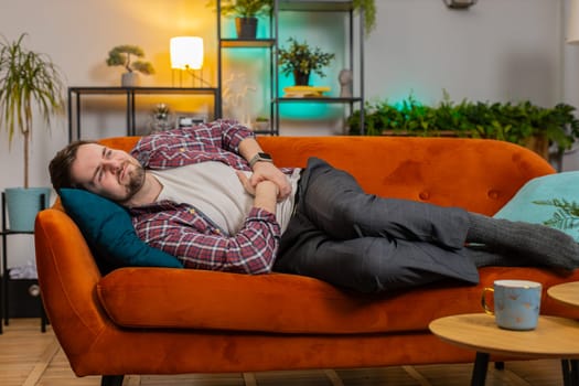 Ill sick young man lying on sofa feeling sudden strong abdominal stomach ache, gastritis problem. Upset guy having symptom poisoning diarrhea indigestion peptic ulcer pancreatitis at home living room