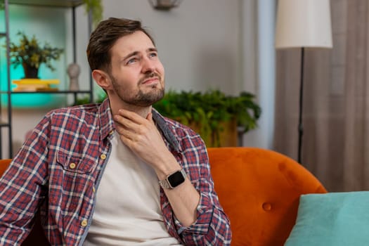 Upset Caucasian ill sick man holds hands over sore throat. Painful neck and frowning, thyroid disorders, suffering sore throat, tonsils inflammation. Young guy sitting on sofa at home room apartments