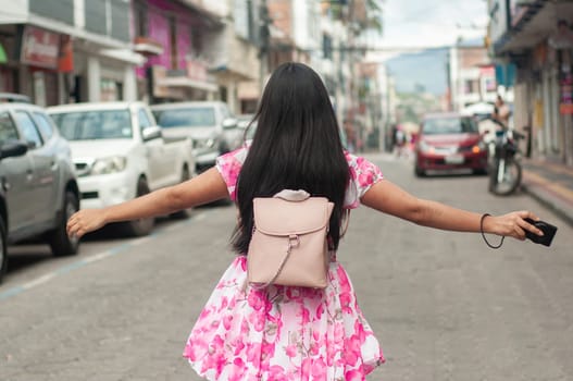 A woman in a pink floral dress with a backpack is seen from behind with her arms spread, walking down an urban street lined with parked cars. High quality photo