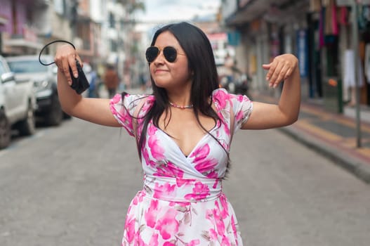 A cheerful young tourist in a pink flowered dress and sunglasses dances in the middle of a busy urban street, arms in the air with a carefree attitude. High quality photo