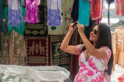 A smiling woman with sunglasses takes a picture amidst the vibrant and colorful textiles of a bustling daytime market. High quality photo
