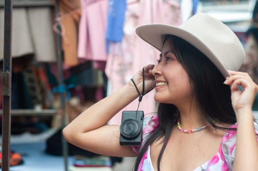 A cheerful young woman with a camera around her neck enjoys her time at a bustling local market, touching the brim of her stylish hat. High quality photo