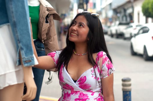 A young tourist in a flowered dress happily amuses herself with the clothes on a mannequin during her shopping trip along a lively street in the city she is visiting on vacation. High quality photo