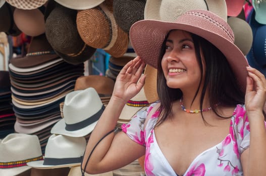 A cheerful woman tries on various hats, surrounded by a colorful selection at an outdoor market stall on a sunny day. High quality photo