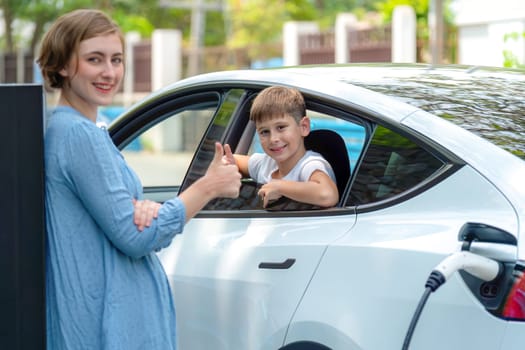 Environmental awareness family with eco-friendly electric car recharging battery from home EV charging station with little boy inside the car. Rechargeable and EV car technology. Perpetual