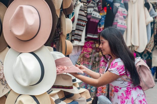 A young, long-haired tourist in a floral print dress carefully chooses a hat from several options at a stall in an open-air market. High quality photo