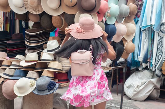 A young girl is seen from behind as she tries on a stylish pink hat amidst a vibrant display of various hats at an outdoor market stall. High quality photo