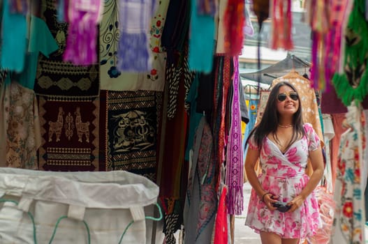 A cheerful woman browses through an array of vibrant textiles hanging at an outdoor marketplace, her sunglasses reflecting the bright daylight. High quality photo