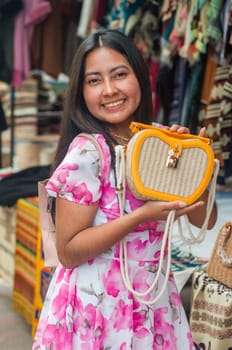 A cheerful young woman in a floral dress holds a stylish handbag while browsing through a colorful outdoor market. High quality photo