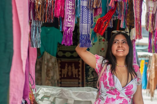 A cheerful woman goes sightseeing and chooses a brightly colored scarf from an assortment of handcrafted fabrics displayed at an open-air market stall. High quality photo