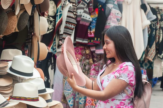 A smiling young woman is selecting a hat from the diverse array displayed at a lively outdoor market stall, surrounded by vibrant textiles. High quality photo