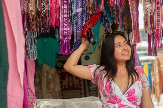 A smiling young woman in a floral dress browses through vibrant, hanging scarves at an outdoor market stall. High quality photo