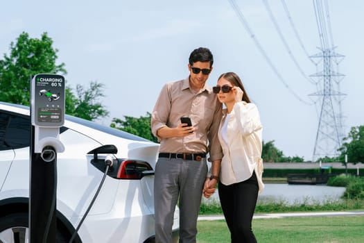 Couple pay for electricity with smartphone while recharge EV car battery at charging station connected to power grid tower electrical as electrical industry for eco friendly car utilization.Expedient