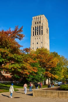 Students stroll under the historic Burton Memorial Tower amid a vibrant autumn setting at the University of Michigan, Ann Arbor, embodying the essence of college life and academia.