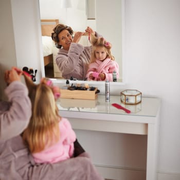 Mom, daughter and playing with hair in mirror for beauty, care and helping or getting ready. Happy mother, family and girl or kid with rollers for hairstyle, fashion and bonding together at home.