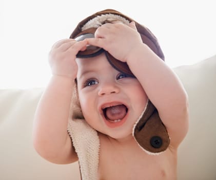 Baby, hat and happy playing or home with laughing for childhood development with curiosity, costume or growing. Kid, accessories and joy in apartment on sofa in Canada or weekend, innocent or cute.