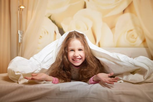 Happy girl lies under a blanket and looking at camera in hotel or in bed room