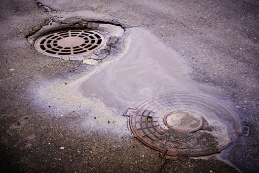 An oil slick against the background of an asphalt road flows into a storm drain against the background of a sewer grate and the lid of a closed well