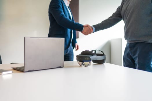 Cropped photo of a business hand shake next to laptop and augmented mixed reality goggles