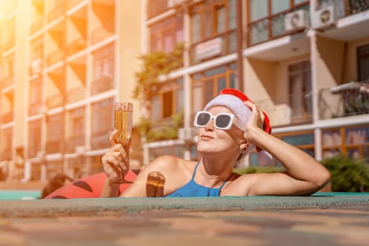 Woman pool Santa hat. A happy woman in a blue bikini, a red and white Santa hat and sunglasses poses near the pool with a glass of champagne standing nearby. Christmas holidays concept