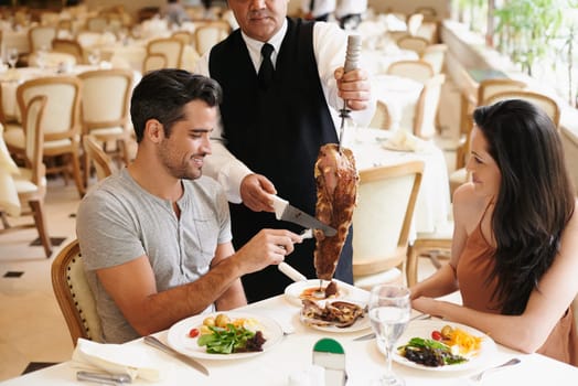 Happy couple, date and dining with kebab of waiter serving, meat or slices on romantic dinner at table. Young man or woman with chef, skewer or food for meal, eating or enjoying service at restaurant.