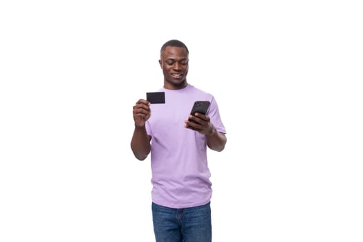 a young joyful slender American student guy dressed in a light lilac T-shirt and jeans uses an online smartphone and a credit card.