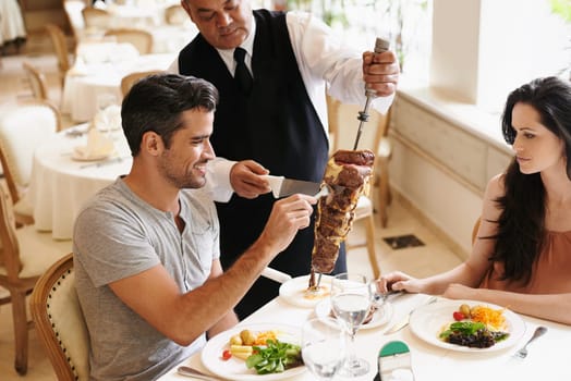 Happy couple, date and waiter with kebab for serving, fine dinning or romantic dinner at table. Young man and woman with chef cutting skewer of food for meal, eating or enjoying service at restaurant.