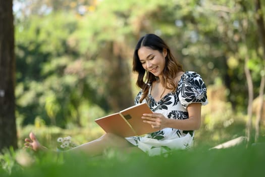 Relaxed young Asian woman reading interesting book and enjoying free time at green park.